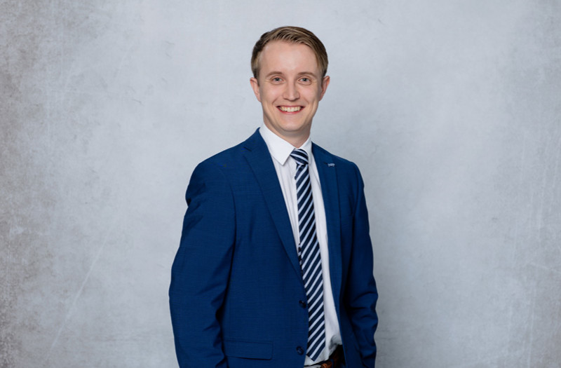 Herr Armin Sielbeck mit blauem Jackett und weißem Hemd steht vor einem hellgrauen Hintergrund und blickt direkt in die Kamera.