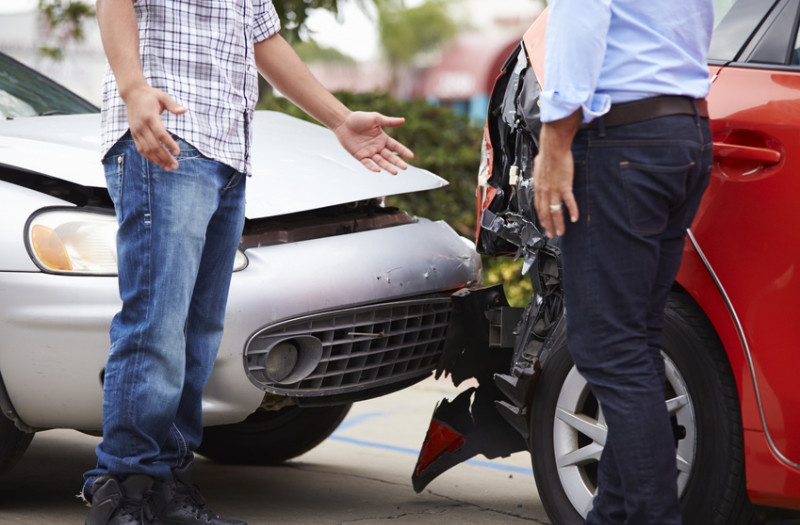 Zwei Personen stehen nach einem Zusammenstoß an der beschädigten Front eines weißen Autos und am Heck eines roten Autos.