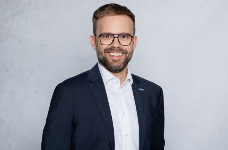 Herr Gunnar Stark mit kurzen Haaren, Bart und Brille, trägt einen dunklen Anzug, ein weißes Hemd und eine hellgraue Krawatte, lächelt in die Kamera.