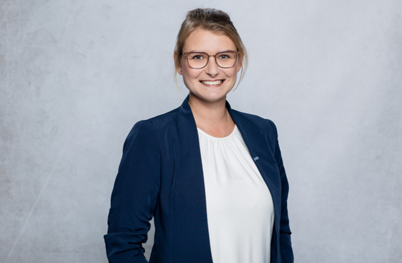 Frau Julia Tiedemann mit zur&uuml;ckgebundenem hellbraunem Haar, Brille, marineblauem Blazer und wei&szlig;er Bluse l&auml;chelt in die Kamera.