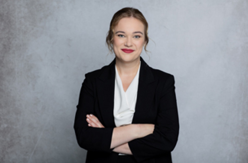 Frau Juliane Rosin mit heller Haut, einem schwarzen Blazer und einer wei&szlig;en Bluse, steht selbstbewusst mit verschr&auml;nkten Armen und l&auml;chelt vor einem Hintergrund.