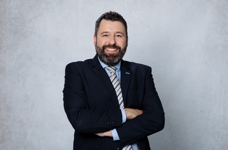 Herr Kai Richtering mit blauem Jackett und hellblauem Hemd steht vor einem schlichten hellgrauen Hintergrund und blickt direkt in die Kamera.