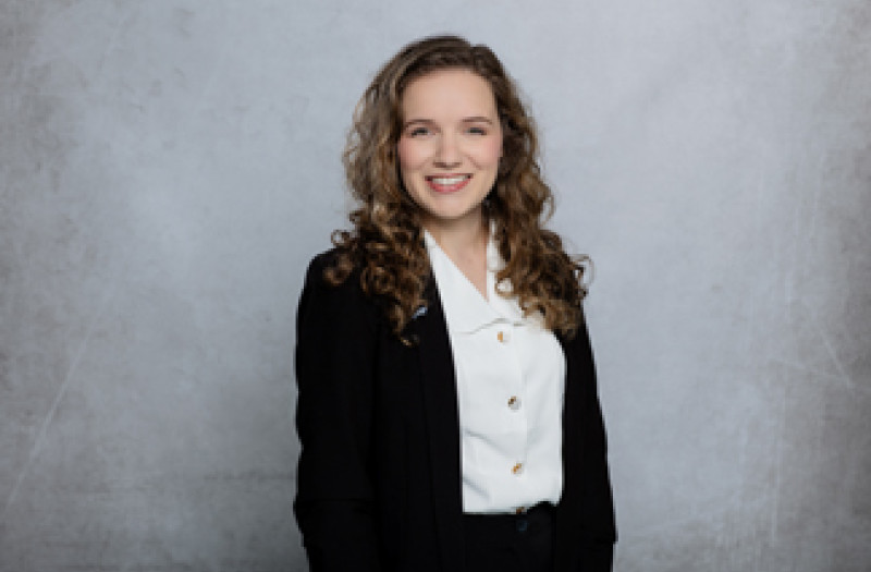 Eine Lara M&uuml;hlenbruch mit lockigem Haar in einem schwarzen Blazer und einer wei&szlig;en Bluse l&auml;chelt in die Kamera.