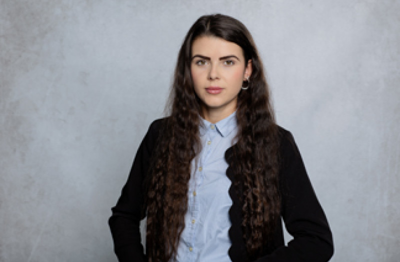 Frau Michelle von Allw&ouml;rden mit langem, welligem braunem Haar, die ein hellblaues Hemd mit Kn&ouml;pfen und eine schwarze Strickjacke tr&auml;gt, steht vor einem hellgrauen Hintergrund.