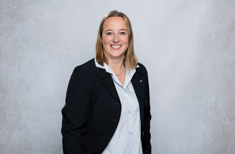 Frau Sandra Reppin mit schwarzer Jacke und hellblauem Hemd steht vor einem hellgrauen Hintergrund und blickt l&auml;chelnd in die Kamera.