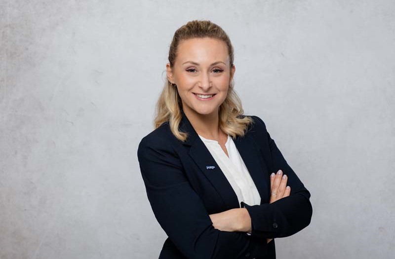 Frau Stefanie Weit mit blauer Jacke und wei&szlig;em Hemd steht vor einem schlichten hellgrauen Hintergrund und blickt direkt in die Kamera.