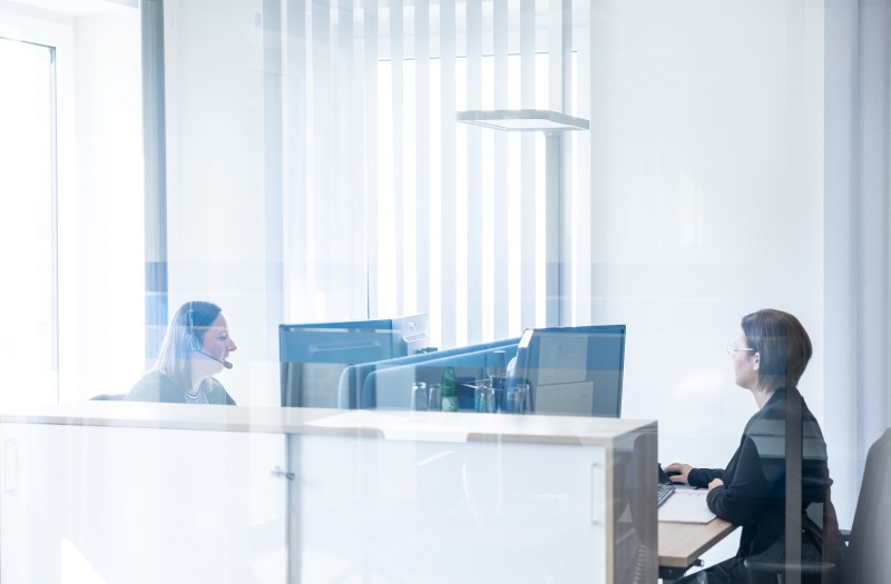 Zwei Frauen arbeiten in einem hellen B&uuml;ro an Schreibtischen, die durch transparentes Glas voneinander getrennt sind.