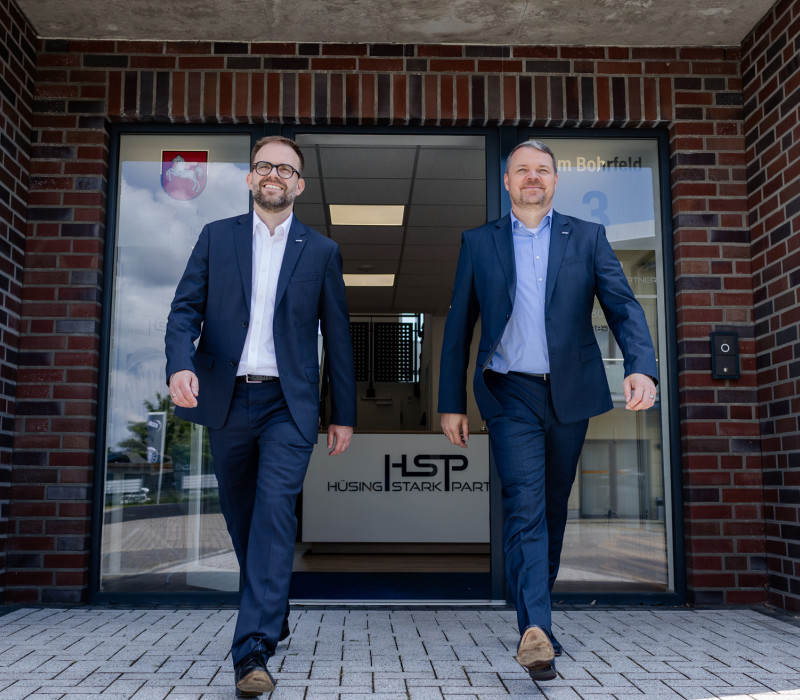 Zwei lächelnde Männer in Anzügen verlassen das Bürogebäude "HSP HÜSING STARK PART".
