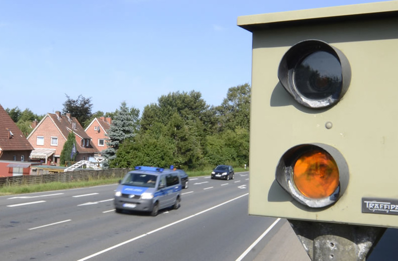 Nahaufnahme einer Radarkamera mit orangefarbenem Licht auf einer mehrspurigen Straße. Mehrere Autos, darunter ein Polizeifahrzeug, fahren vorbei.