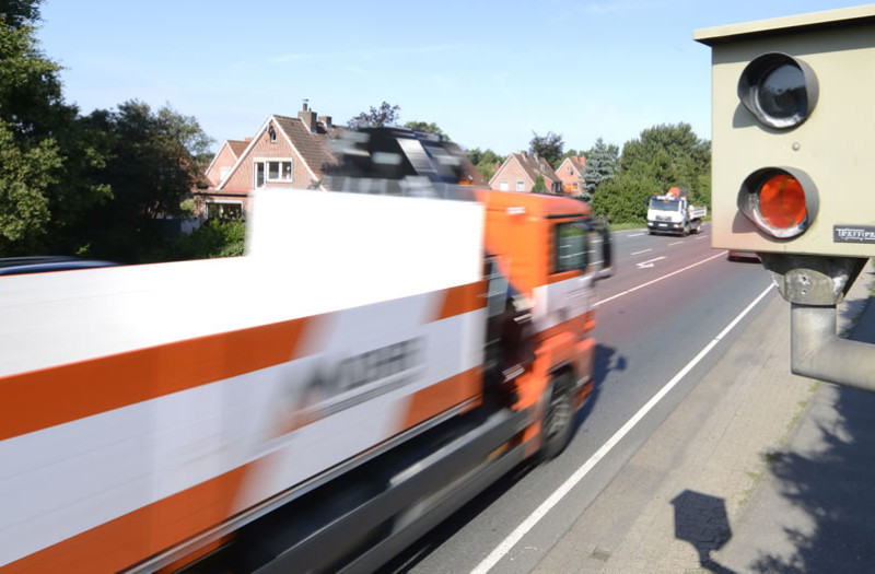Ein schnell fahrender LKW wird von einer Radarkamera am Straßenrand in Bewegungsunschärfe auf einer Vorstadtstraße mit Häusern und Bäumen im Hintergrund erfasst.