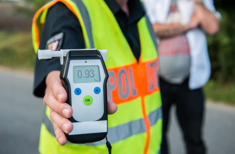 Ein Polizist in einer Neonweste hält ein Alkoholtestgerät mit einem Wert von 0,93 in der Hand, während im Hintergrund eine weitere Person zu sehen ist.
