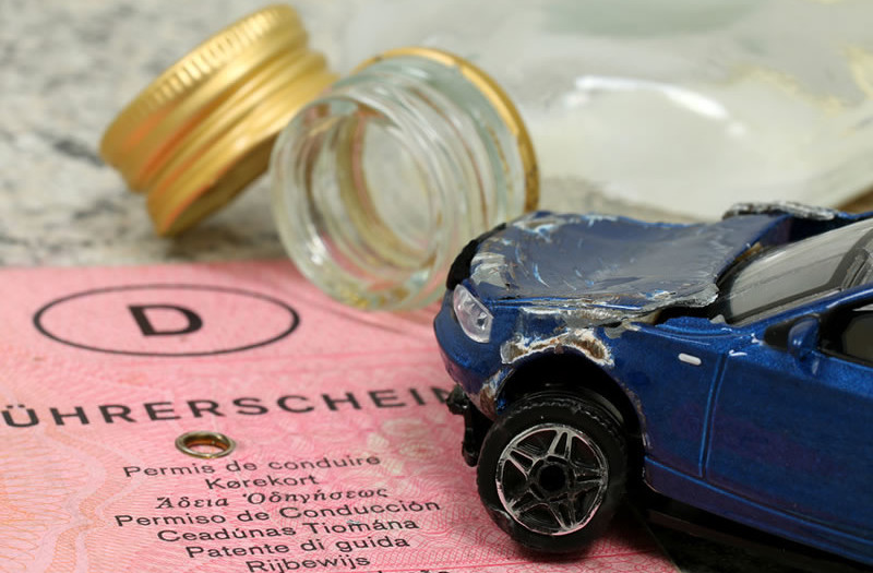 Ein Spielzeugauto mit Unfallschaden liegt auf einem deutschen Führerschein. Im Hintergrund sind ein offenes Glas und eine verschüttete Flüssigkeit zu sehen.