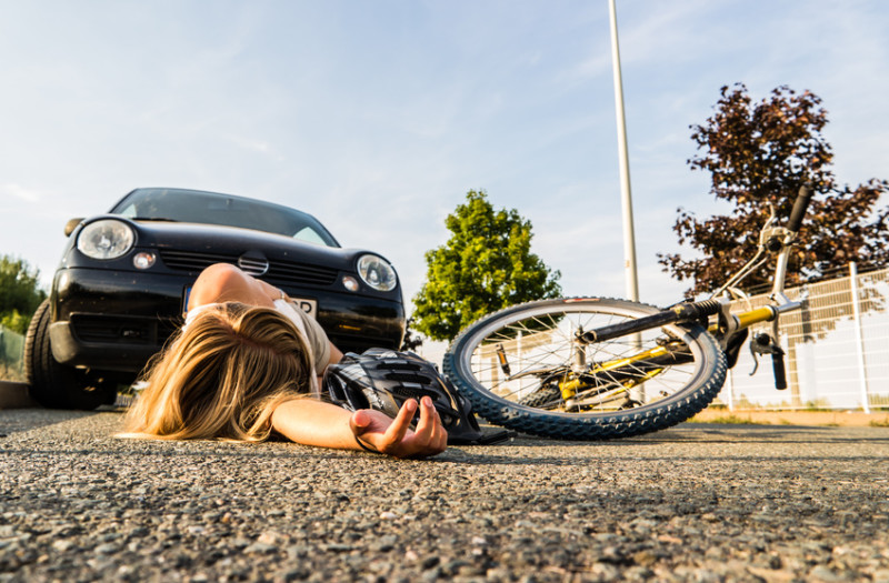 Eine Person liegt neben einem umgestürzten Fahrrad vor einem Auto auf dem Boden, was auf einen möglichen Unfall hindeutet.