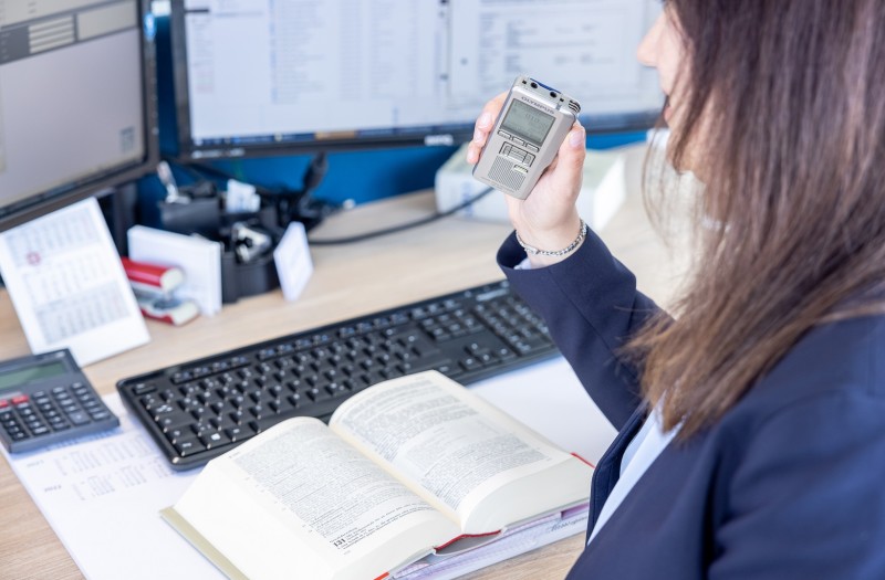 Eine Frau im Business-Outfit sitzt mit offenem Buch und Diktiergerät an einem Schreibtisch voller Büromaterial, Tastatur und Taschenrechner.