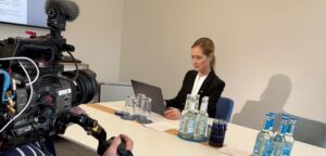 Sabrina Stark sitzt vor ihrem Laptop am Tisch. Auf sie ist eine Fernsehkamera gerichtet.