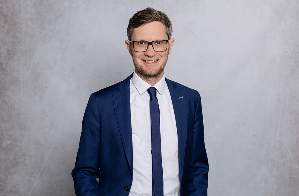 Herr Christian Stahlich mit Brille, blauen Jackett und weißem Hemd steht vor einem schlichten hellgrauen Hintergrund.