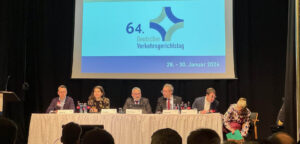 Sechs Personen sitzen auf dem Podium an einem Tisch. Im Hintergrund sind auf der Leinwand das Logo und der Schriftzug des 64. Deutschen Verkehrsgerichtstages zu sehen.