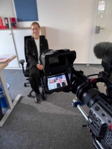 Sabrina Stark lächelt in eine Fernsehkamera, die sich im Vordergrund befindet. Auf dem Bildschirm der Kamera ist der Ausschnitt der Aufnahme zu sehen.