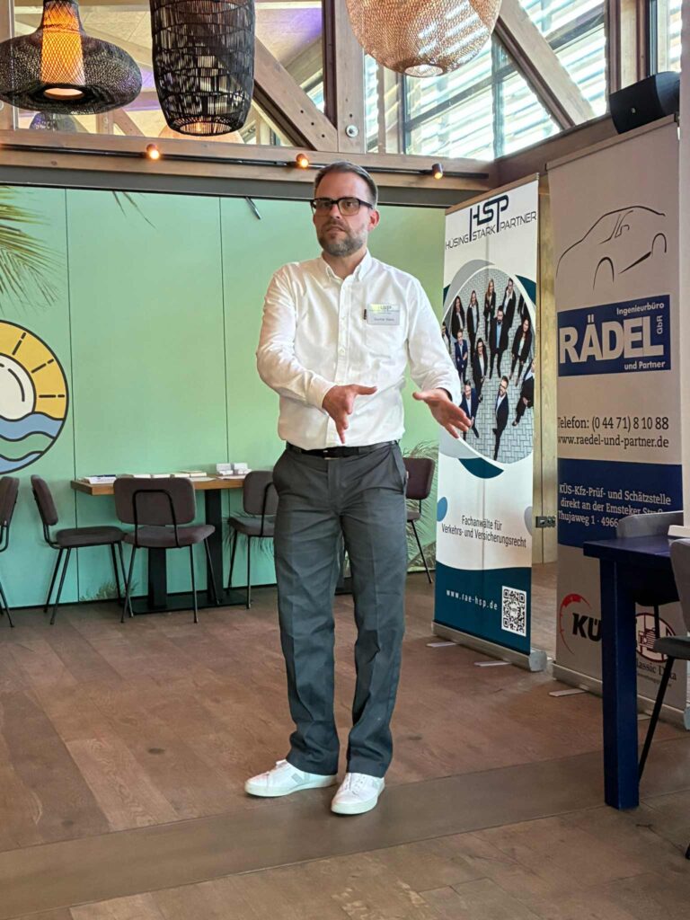 Ein Mann mit Brille, weißem Hemd und grauer Hose gestikuliert in einem Saal. Im Hintergrund sind Werbebanner.